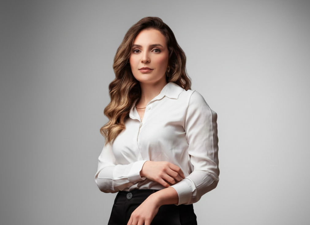 Pessoa com cabelo castanho ondulado e longo, vestindo camisa branca e calça preta, em pé contra fundo cinza neutro, com pose formal e expressão serena.